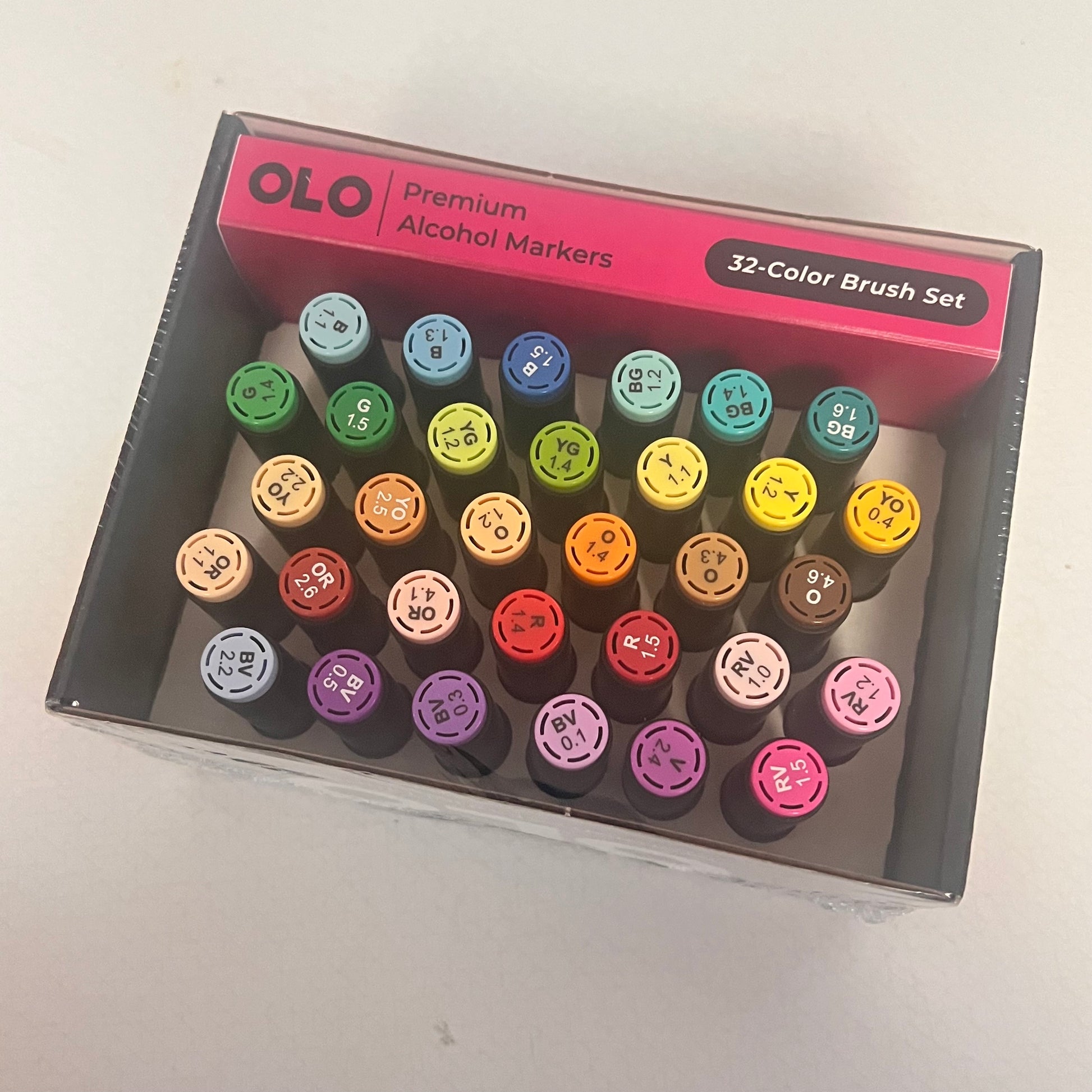 A 32-color brush set of OLO premium alcohol markers arranged inside their pink and gray retail box, photographed from above on a light surface.